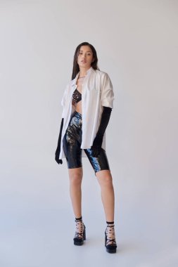 Kişisel stil, esmer saçlı, gri arka planda poz veren genç Asyalı kadın, beyaz gömlek ve siyah eldiven, lateks şortla poz veren manken, cesur tarz, kenar moda, tam boy
