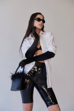 Cesur stil, moda anlayışı, güneş gözlüklü Asyalı kadın gri arka planda tüylü el çantalı poz, lateks şortlu genç model, siyah eldiven ve beyaz gömlek, kavramsal gömlek. 