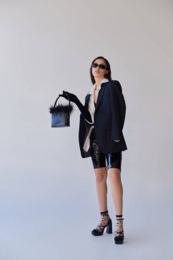 Moda anlayışı, moda anlayışı, güneş gözlüklü esmer Asyalı kadın gri arka planda tüylü çantalı poz, lateks şortlu model, siyah ceket ve eldiven, tam boy 