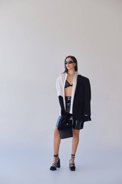 Havalı bir stil, stüdyo fotoğrafçılığı, şık görünümlü genç Asyalı kadın ve gri arka planda tüylü el çantasıyla poz veren güneş gözlüğü, spor ceket ve lateks şort, gençlik modası, tam boy. 