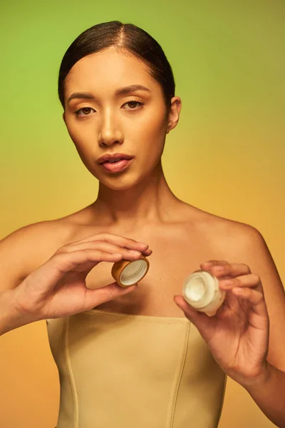 美容品 年轻的亚洲女人 光着肩膀 拿着面霜的化妆品罐 看着绿色背景的相机 美容业 光彩夺目的皮肤 — 图库照片