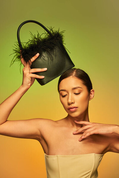 beauty and style, brunette asian woman with bare shoulders posing with feather purse on green background, hand near face, gradient, fashion statement, glowing skin, natural beauty, young model 