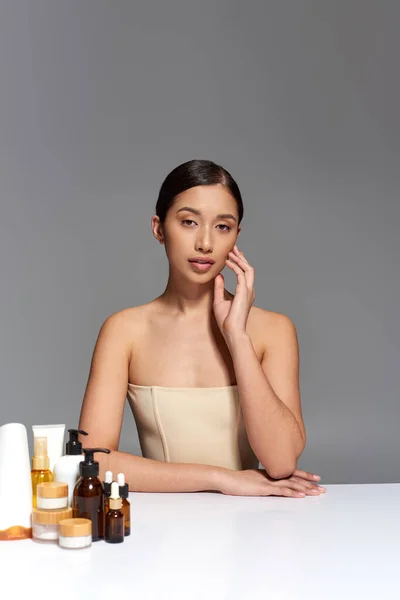 プロダクトプレゼンテーション スキンケア 黒髪と若いアジアのモデルは グレーの背景に異なる美容製品の近くにポーズ 輝くと健康肌 美容キャンペーン 顔の治療の概念 — ストック写真