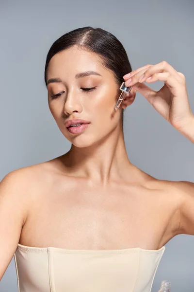 プロダクトプレゼンテーション スキンケア ブルネットヘアで若いアジアのモデルグレーの背景にピペットで血清を適用 輝くと健康的な肌 美容キャンペーン 顔の治療の概念 — ストック写真