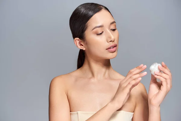 プロダクトプレゼンテーション スキンケア ブロンドの髪を持つ若いアジアのモデルは 灰色の背景にクリームと化粧瓶を保持します 輝くと健康肌 美容キャンペーン 顔の治療コンセプト — ストック写真