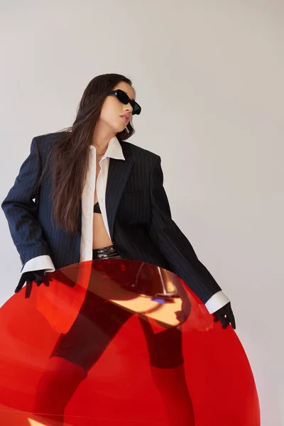 采购产品现代时尚 工作室摄影 年轻的亚洲女人在时髦的外观和太阳镜摆出接近红色圆形玻璃 灰色背景 夹克和乳胶短裤 年轻的时尚 凉爽的风格 — 图库照片