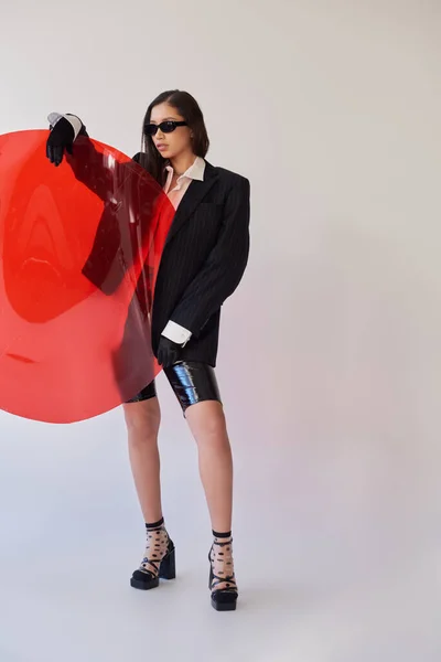 漂亮的亚洲女人 长相时尚 太阳镜造型 手持红色圆形玻璃 灰色背景 夹克和乳胶短裤 年轻模特 时尚前卫 工作室摄影 — 图库照片