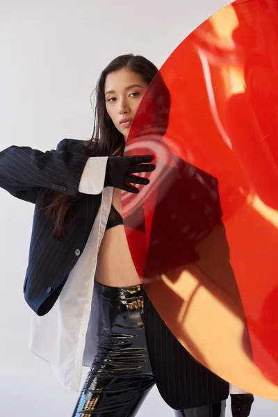 漂亮的亚洲女人 身穿时髦服装 手持红色圆形玻璃杯 灰色背景 夹克和乳胶短裤 年轻模特 时尚前卫 摄影棚摄影 概念设计 — 图库照片