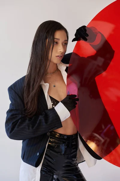 黑发亚洲女人 身穿时髦服装 手持红色圆形玻璃杯 灰色背景 夹克和黑色乳胶短裤 戴着手套的年轻模特 时尚潮流 工作室摄影 — 图库照片