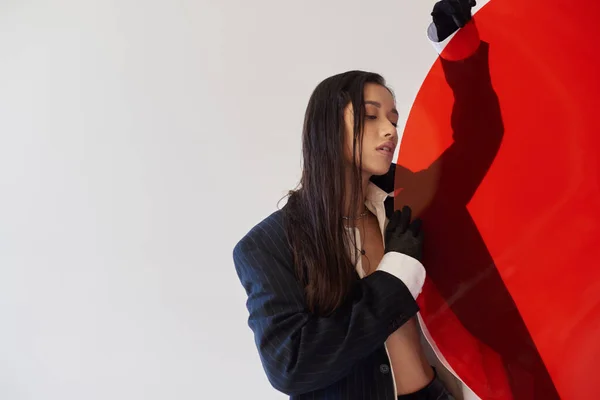 赤い丸い形のガラス グレーの背景 ブレザーと黒のラテックスのショートパンツ 手袋で若いモデルを保持流行の服でかなりアジアの女性 ファッションの選択 スタジオの写真 — ストック写真