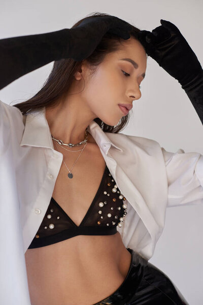 beauty and fashion, perfect skin, young asian woman posing with raised hands on grey background, black gloves and white shirt, bra look, fashion statement, conceptual, personal style