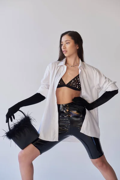 エッジスタイル ファッションステートメント グレーの背景に羽のハンドバッグを身に着けているアジアの女性 離れて見て若いモデル 黒手袋と白のシャツ ラテックススタイル 概念的 個人的なスタイル — ストック写真