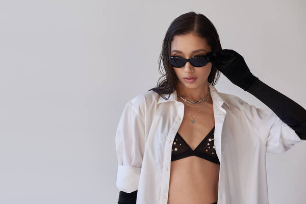 бюстгальтер взгляд, мода заявление, стильная азиатская женщина в солнцезащитных очках позирует на сером фоне, молодая модель в черных перчатках и белой рубашке, идеальная кожа, концептуальный, модный наряд, молодой  