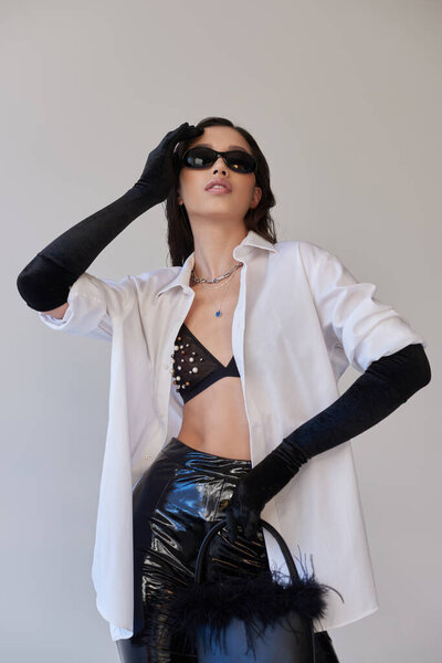 trendy look, fashion statement, brunette asian woman in sunglasses posing with feathered purse on grey background, young model in latex shorts, black gloves and white shirt, conceptual 