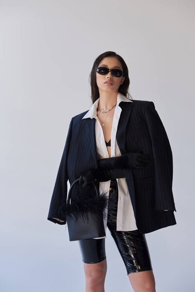 personal style, brunette asian woman in dark sunglasses posing with feathered handbag on grey background, young model in latex shorts, bra, blazer and black gloves, style and trends 