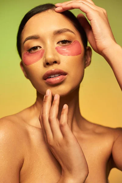 pretty and brunette asian woman posing with eye patches on green background, gradient, beauty, detox skin, facial treatment, bare shoulders, skincare routine, glowing skin, portrait