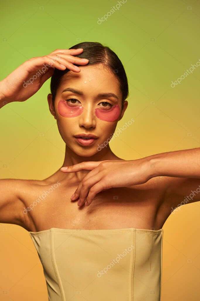 Skincare campaign, young asian woman with brunette hair and clean skin posing and looking at camera on green background, bare shoulders, moisturizing eye patches, glowing skin