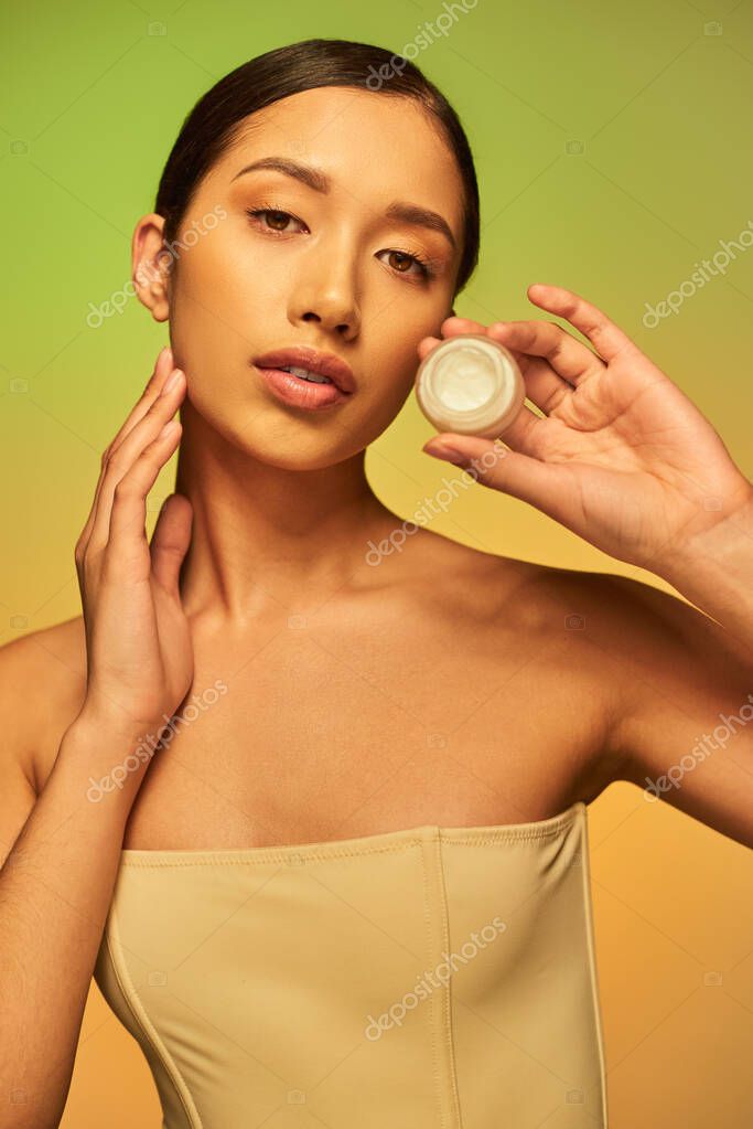 Beauty product, young asian woman with bare shoulders holding cosmetic jar with face cream on green background, brunette hair, beauty industry, glowing skin, skin care concept