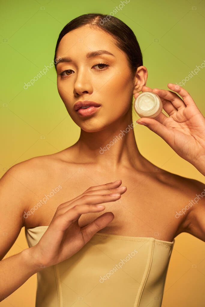 Beauty photography, young asian woman with bare shoulders holding cosmetic jar with cream on green background, brunette hair, beauty industry, glowing skin, skin care concept