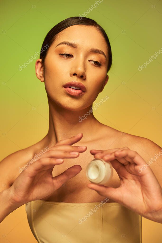 Beauty campaign, young asian woman with bare shoulders holding cosmetic jar with face cream on green background, brunette hair, beauty industry, glowing skin, skin care concept