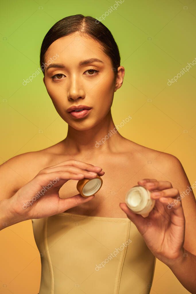 Beauty product, young asian woman with bare shoulders holding cosmetic jar with face cream and looking at camera on green background, brunette hair, beauty industry, glowing skin