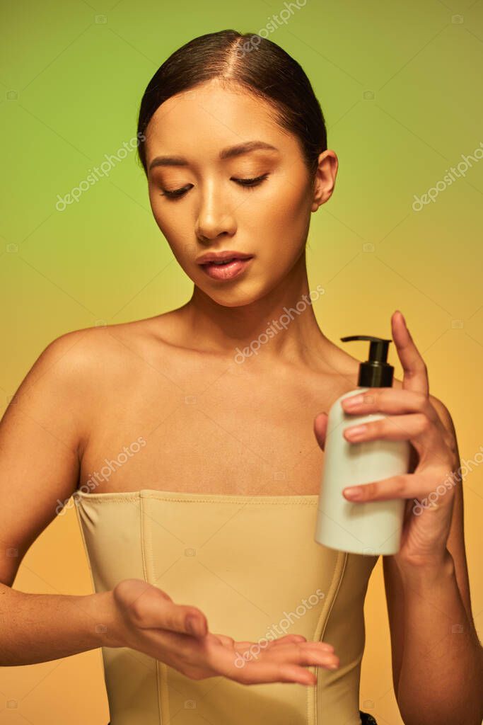 Product presentation, skin care product, young asian woman with bare shoulders holding cosmetic bottle and posing on green background, glowing skin, brunette hair