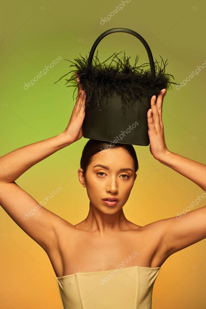 Beauty and style, brunette asian woman with bare shoulders posing with feather purse on head on green background, gradient, fashion statement, glowing skin, natural beauty, young model