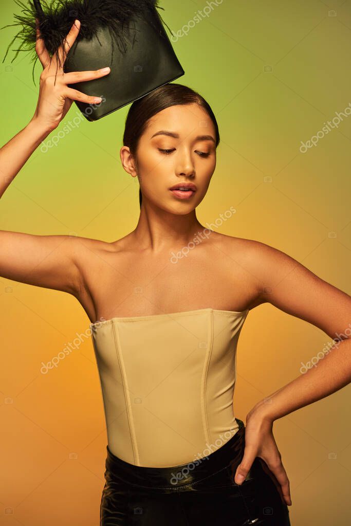 Beauty and style, brunette asian woman with bare shoulders posing with feather purse and hand on hip on green background, gradient, fashion statement, glowing skin, natural beauty, young model