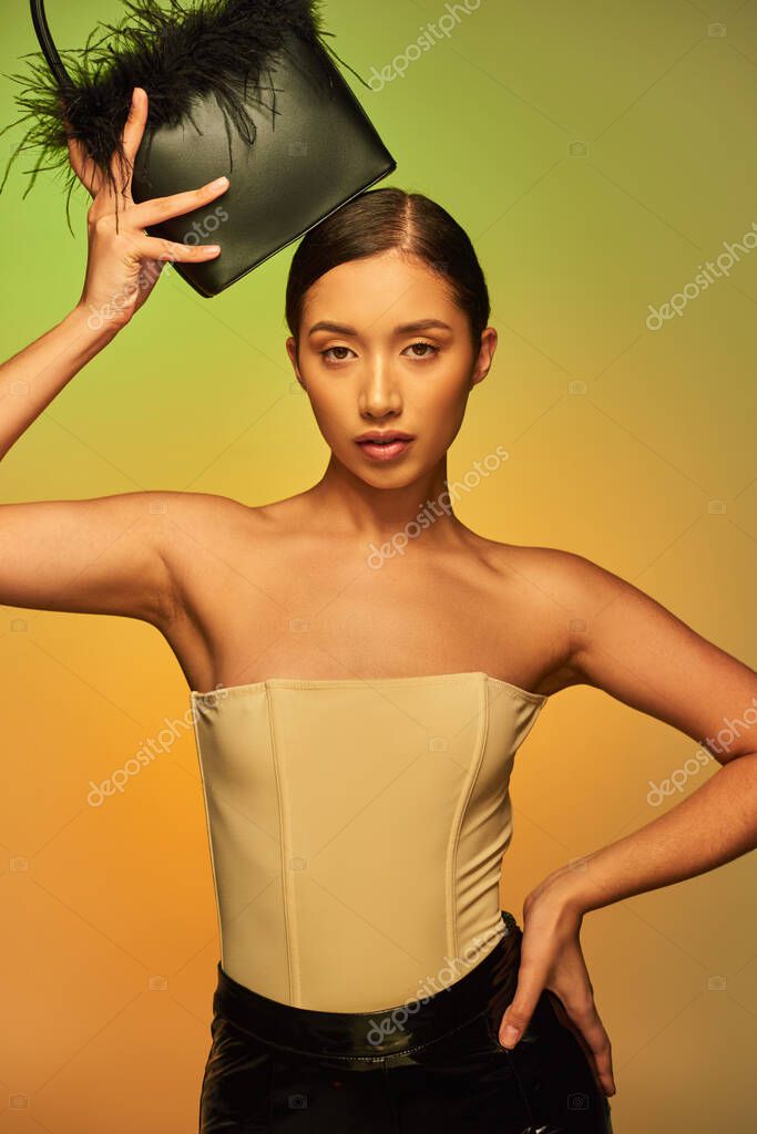 Fashion choices, brunette asian woman with bare shoulders posing with feather purse and hand on hip on green background, gradient, fashion statement, glowing skin, natural beauty, young model