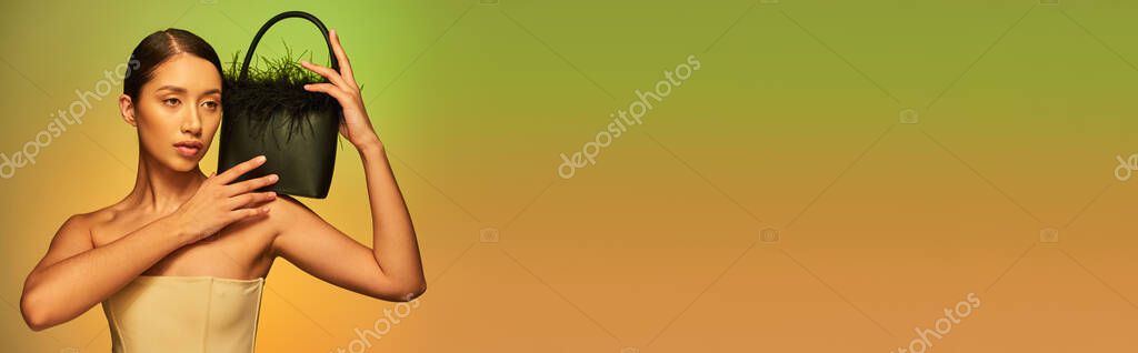 Fashion choices, brunette asian woman with bare shoulders posing with feather purse on green background, gradient, fashion statement, glowing skin, natural beauty, young model, banner