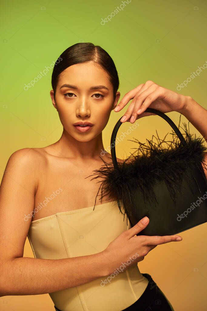 Fashion forward, brunette asian woman with bare shoulders posing with feather purse on green background, gradient, fashion statement, glowing skin, natural beauty, young model looking at camera