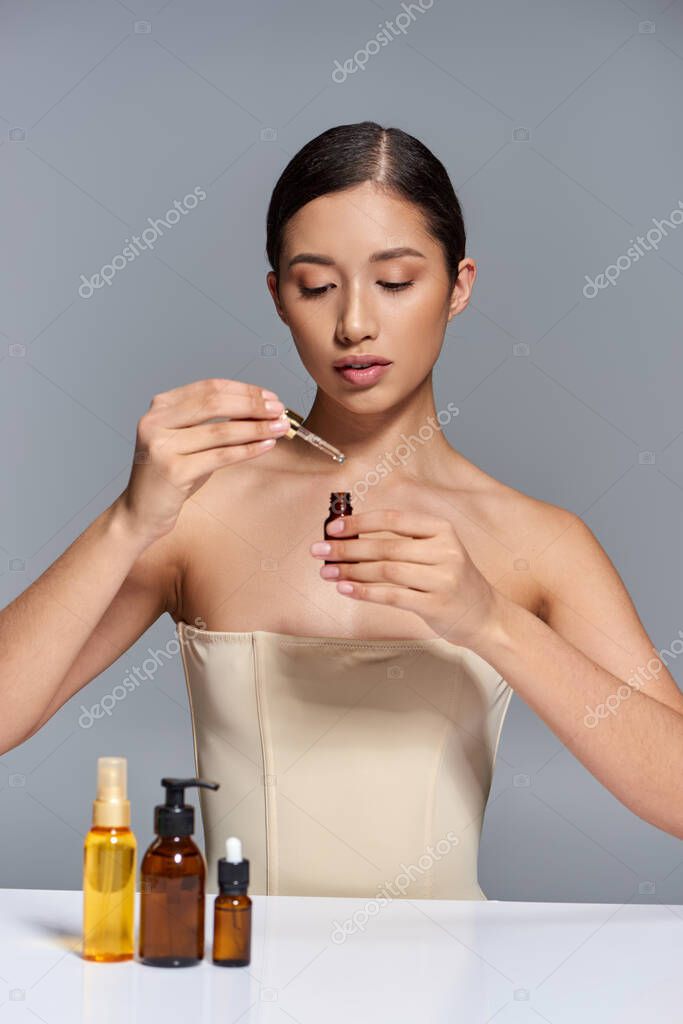 Product presentation, skin care, young asian model with brunette hair holding pipette with oil on grey background, glowing and heathy skin, beauty campaign, facial treatment concept
