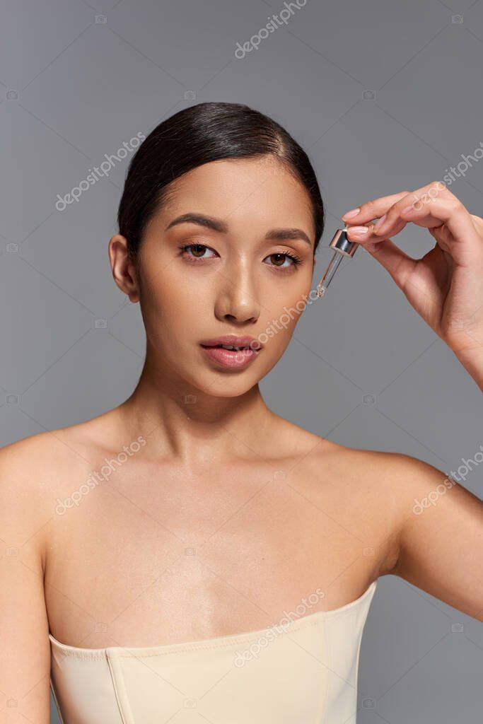 Skin care, young asian woman with brunette hair applying serum with pipette on grey background, glowing and heathy skin, beauty campaign, facial treatment concept, looking at camera