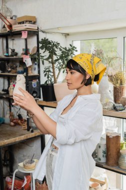 Young brunette asian artist in headscarf and workwear holding clay product while standing in blurred pottery class at background, pottery studio with artisan at work clipart