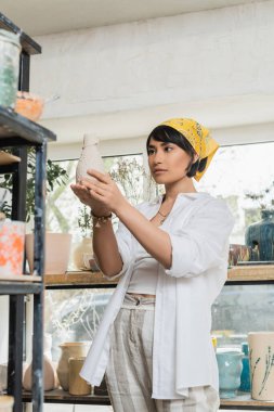 Brunette asian female artist in headscarf and workwear holding clay product while standing near shelves in blurred pottery studio, pottery studio with artisan at work clipart