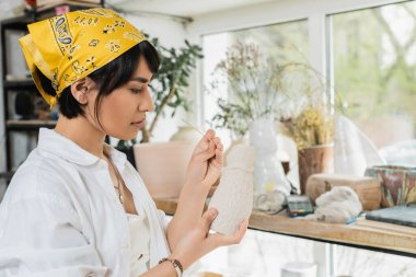 Young asian brunette artist in headscarf and workwear holding clay sculpture and wooden stick while working in blurred pottery class at background, pottery studio with artisan at work clipart