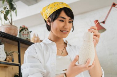 Smiling young asian female artist in headscarf and workwear holding wooden stick and clay sculpture while working in blurred pottery class, pottery studio with artisan at work clipart