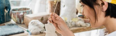 Arka planda bulanık çanak çömlek kursunda çalışırken elinde tahta sopa ve kil heykel tutan Asyalı genç sanatçının yan görüntüsü, zanaatkar ile çömlek stüdyosu, afiş. 