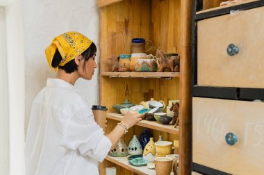 Side view of young asian female artist in headscarf and workwear holding coffee to go while standing near clay products on shelves in pottery workshop, pottery workshop with skilled artisan clipart