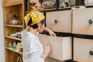 Side view of young asian female artist in headscarf and workwear opening cupboard while working in blurred pottery workshop, pottery workshop with skilled artisan clipart