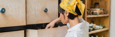 Side view of young asian female artist in headscarf opening cupboard while standing in blurred pottery workshop at background, pottery workshop with skilled artisan, banner  clipart