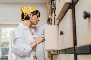 Side view of young brunette asian female artist in workwear and headscarf opening cupboard while working in blurred pottery class, pottery workshop with skilled artisan clipart