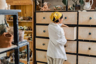 Side view of young brunette asian craftswoman in headscarf and workwear opening cupboard while standing and working in ceramic studio, pottery workshop with skilled artisan clipart