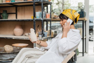 Side view of young asian artist in headscarf and workwear talking on smartphone and holding ceramic sculpture while sitting in ceramic workshop, artisan in pottery studio focusing on creation clipart