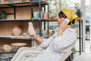 Genç esmer Asyalı kadın çömlekçi başörtüsü ve iş kıyafetleriyle akıllı telefondan konuşuyor ve seramik atölyede otururken kil heykel tutuyor, çömlek atölyesinde zanaatkar yaratmaya odaklanıyor.