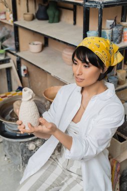 Young asian female potter in headscarf and workwear holding clay product while sitting and working in ceramic workshop at background, craftsmanship in pottery making clipart
