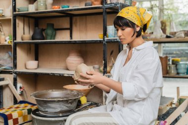 Arka plandaki seramik atölyesinde çömlek çarkının yanında çalışırken başörtüsü ve atkı taşıyan genç Asyalı kadın zanaatkar çömlekçilik yapıyor.
