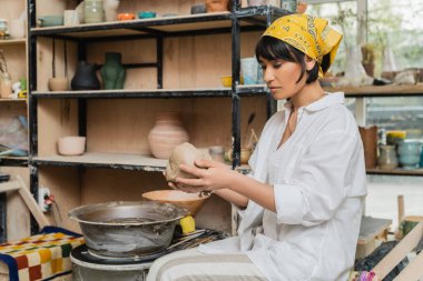Young asian female artisan in headscarf and workwear holding clay while working near pottery wheel in ceramic workshop at background, craftsmanship in pottery making clipart
