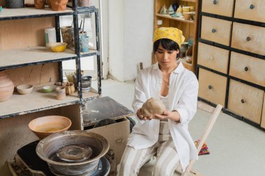 Young brunette asian female potter in workwear and headscarf holding clay near pottery wheel while working in blurred ceramic workshop, craftsmanship in pottery making clipart