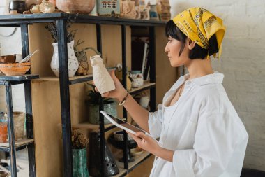 Side view of young asian female artisan in headscarf and workwear holding digital tablet with blank screen and clay sculpture and standing in ceramic workshop, creative process of pottery making clipart